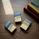 Three rectangular handmade soap bars with a blue and cream marbled pattern are arranged on a dark wood tabletop next to a gold metal divider and a stack of books.
